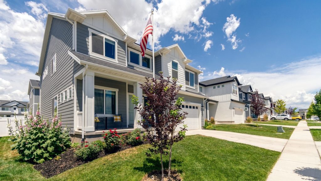 Wide-angle view of a gray two-story suburban home with white trim, an attached garage, an American flag, and a landscaped front yard on a sunny, partly cloudy day.